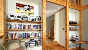 A cozy living room with wooden bookshelves filled with books, a green armchair, and a painting of a racing car. - Large home Assoc Beach across street Firepit Scrnd Porches (Union Pier)