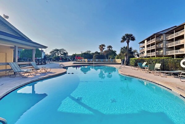 A clear swimming pool with lounge chairs and a building with balconies in the background. - Lighthouse Point 23C | Beachfront 270° View | Top Floor Corner Oasis | 3 Pools (Tybee Island)