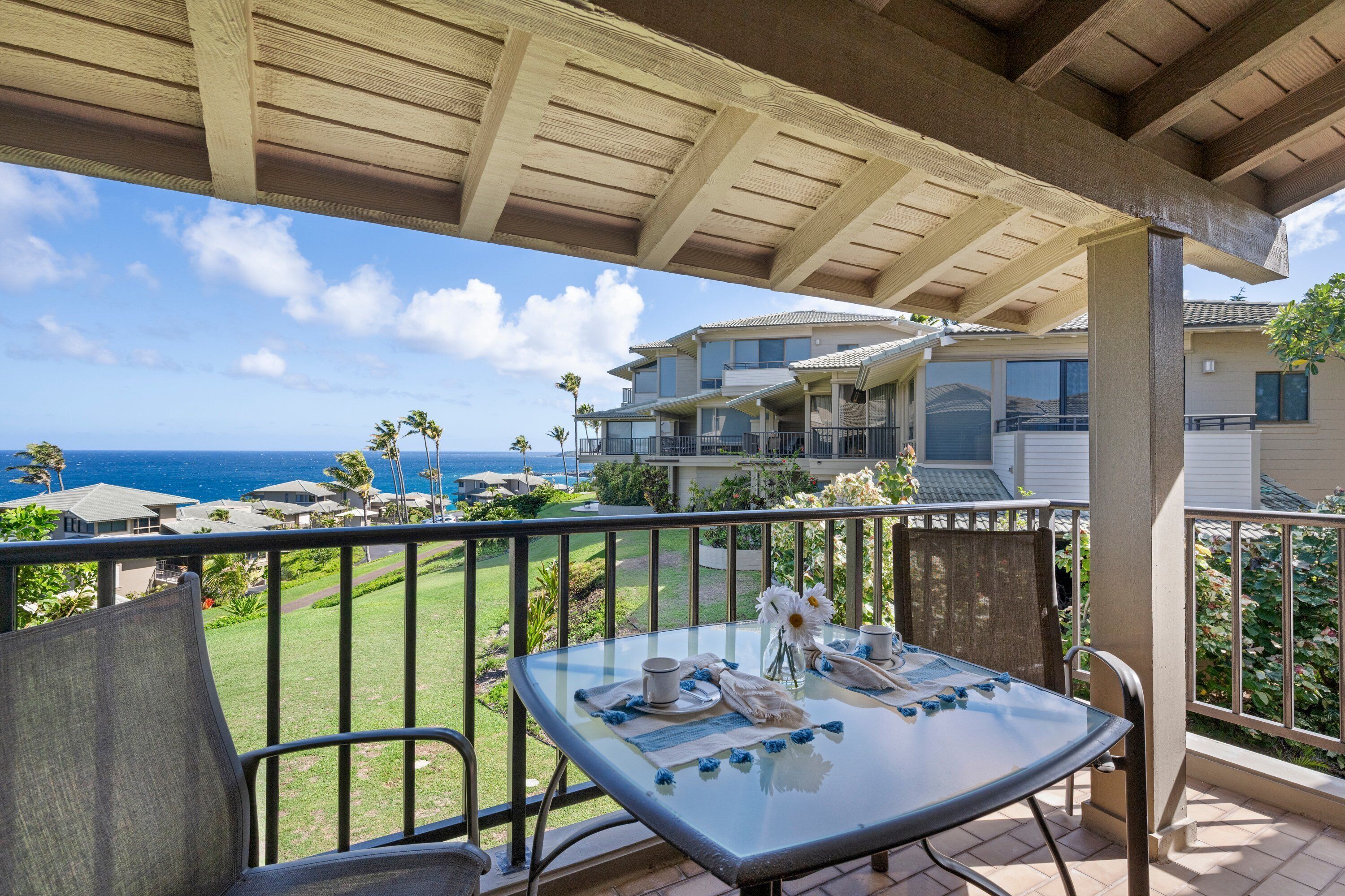 A balcony with a glass table set for dining, overlooking a residential area with houses and a view of the ocean.