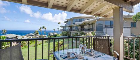 A balcony with a glass table set for dining, overlooking a residential area with houses and a view of the ocean.