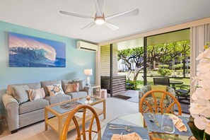 A modern living room with a large ocean wave painting, a ceiling fan, and a view of a tropical garden through large windows.
