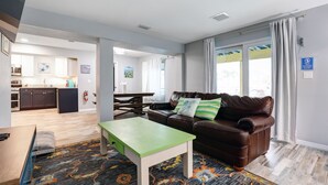 Ein modernes Wohnzimmer mit einer braunen Ledersofa, einem grünen Couchtisch und einem gemusterten Teppich. Im Hintergrund ist eine Küche mit weißen Schränken und einem Essbereich erkennbar.