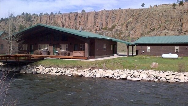 A lodge with a large deck and a covered patio, situated by a river with a rocky shoreline.