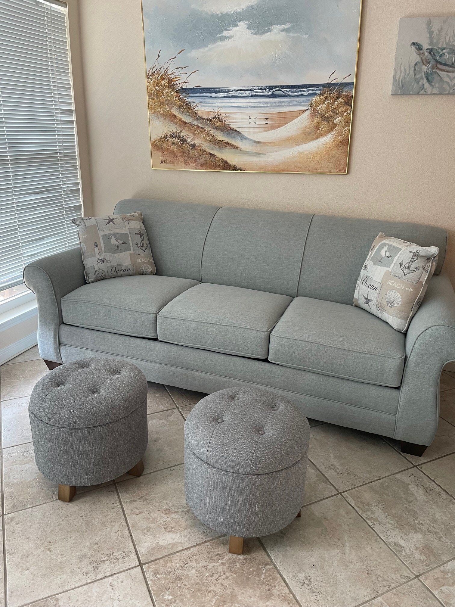 A grey sofa with matching ottomans and decorative pillows, a beach-themed painting above, and a window with blinds.