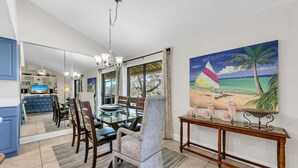 A dining area with a glass table and chairs, a wooden console table with decorative items, and a beach-themed painting on the wall.