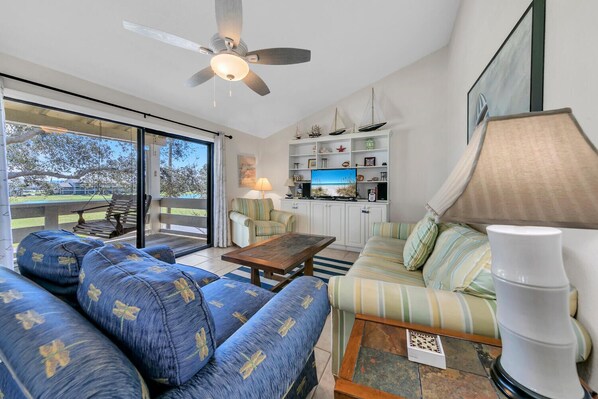A spacious living area with a sofa, a coffee table, and a television. There is a ceiling fan, a lamp, and a bookshelf with various items. - Golf Course 6D - Serene golf course villa just steps to the beach and pools! (Miramar Beach)