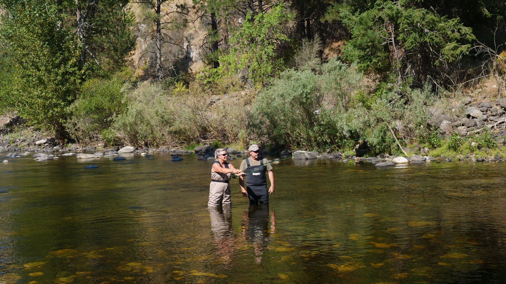Two people standing in a river surrounded by greenery and rocks.