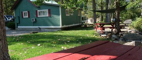 A green cabin with a red roof, surrounded by trees and a picnic table.