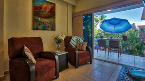 A room with two red armchairs, a small table with a vase of flowers, and a painting on the wall. There is a balcony with a blue umbrella and outdoor furniture.