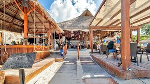 En restaurang vid stranden med träkonstruktioner, en bardel och utsikt över stranden och bergen.