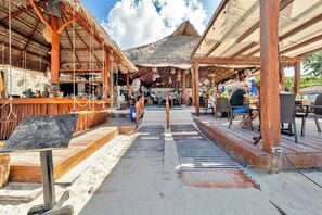 Un restaurant situé en bord de mer, doté de structures en bois, d’un espace bar et offrant une vue sur la plage et les montagnes.