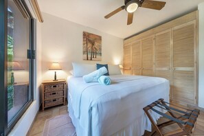 A bedroom with a bed, bedside lamps, a wooden wardrobe, a ceiling fan, and a window with a view.