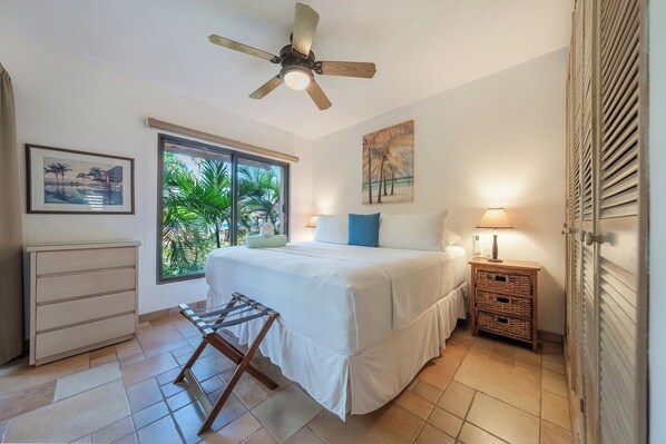 A bedroom with a bed, a nightstand, a ceiling fan, a window with a view of greenery, and a door with a louvered design.