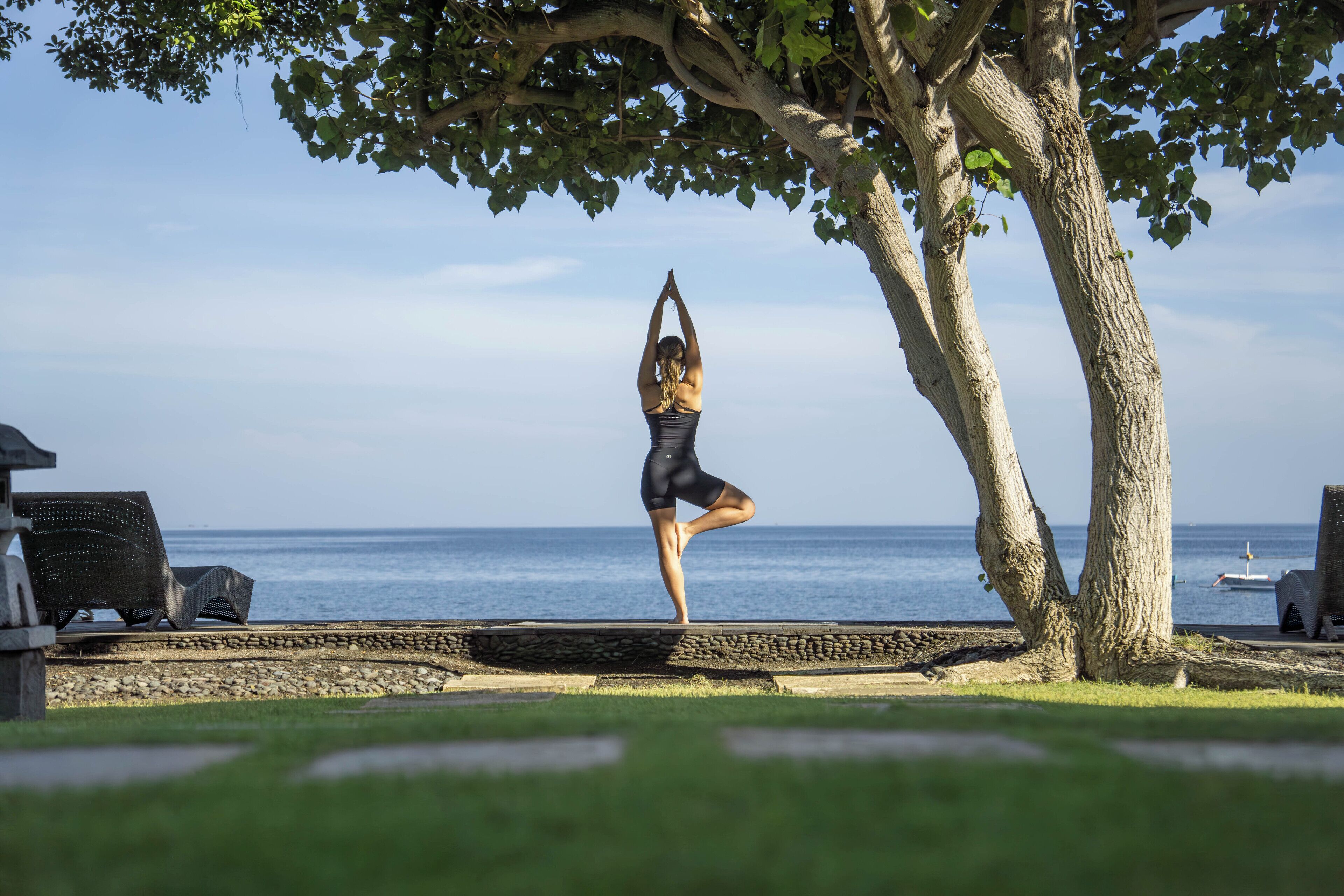 Iemand doet een yoga-houding op een stenen platform aan zee, met een boom en een stoel op de voorgrond.