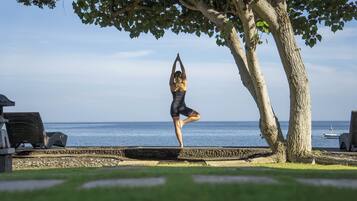 Iemand doet een yoga-houding op een stenen platform aan zee, met een boom en een stoel op de voorgrond.