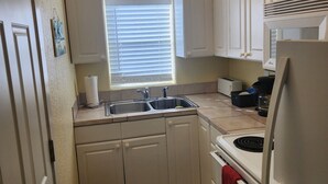 Una cocina con armarios blancos, un lavamanos, una estufa y un refrigerador. Hay una ventana con persianas, un cubo de basura y una planta en maceta sobre la encimera.