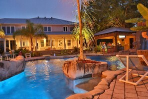 A pool area with a stone planter, lounge chairs, and a building with a covered patio in the background. - Private Resort-Style Pool w/ Lazy River, Grotto & Hot Tub – Golfers & Pups Love! (Lutz)