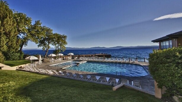 A pool area with lounge chairs and umbrellas, a wooden deck, and a view of the sea and mountains.