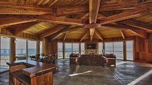 A spacious room with a high ceiling, wooden beams, a bar area, and large windows offering a view of the ocean.