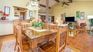 A spacious dining area with a glass-top table surrounded by wooden chairs, a ceiling fan, and a television in the background. - Nihi Kai 422 Ocean Views, 5 Minute Walk to Beach, Five Stars. Booking Specials! (Koloa)