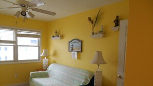 Una habitación con pared amarilla, una cama con colcha a rayas, un ventilador de techo, una ventana con persianas y un estante de pared con objetos decorativos.