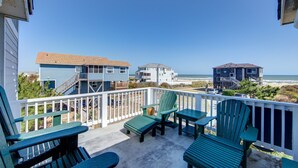 Ein Balkon mit grünen Adirondack-Stühlen und einem Tisch mit Blick auf einen Strand, im Hintergrund Häuser.