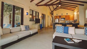 Amplia sala de estar con un conjunto de sofás, una mesa de centro y una televisión. Se observa una zona de cocina con armarios de madera y una mesa de comedor.