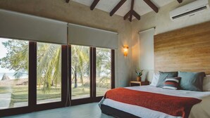 A bedroom with a large bed, a nightstand, a wall-mounted lamp, and a view of palm trees and beach through floor-to-ceiling windows. - Beachfront home + private pool, Playa Venao, Sleeps 10+ (Playa Venao)