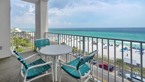 A balcony with a table and chairs, overlooking a beach with parked cars and umbrellas.