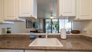 A kitchen with granite countertops, white cabinets, a built-in sink, a ceiling fan, and a view of the outdoors. - Maui Condo Ocean and Park Views Kealia Unit 205 Tranquil Beach Front Resort (Kihei)