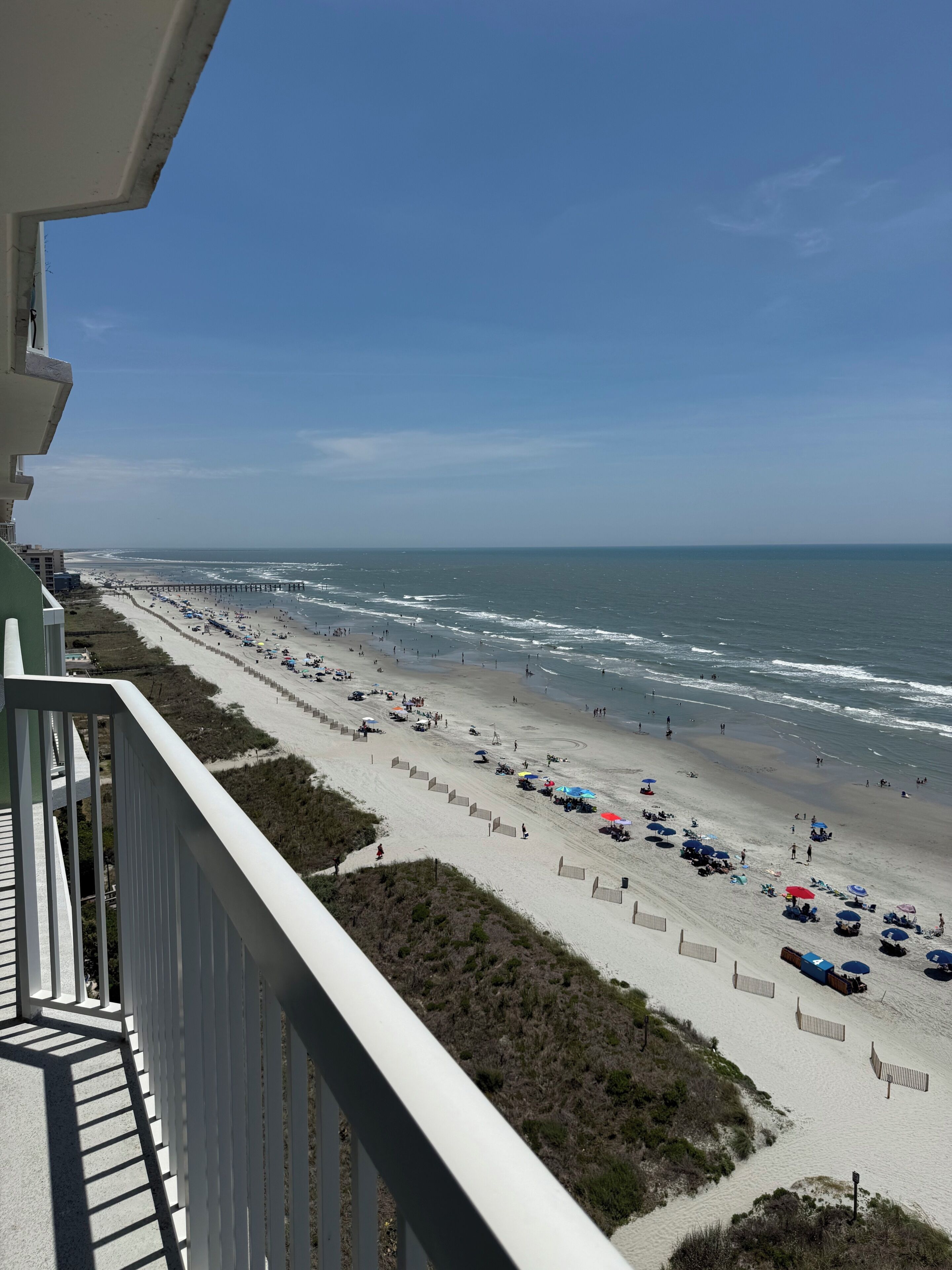 A beach with umbrellas and people, a pier in the distance, and a clear sky.