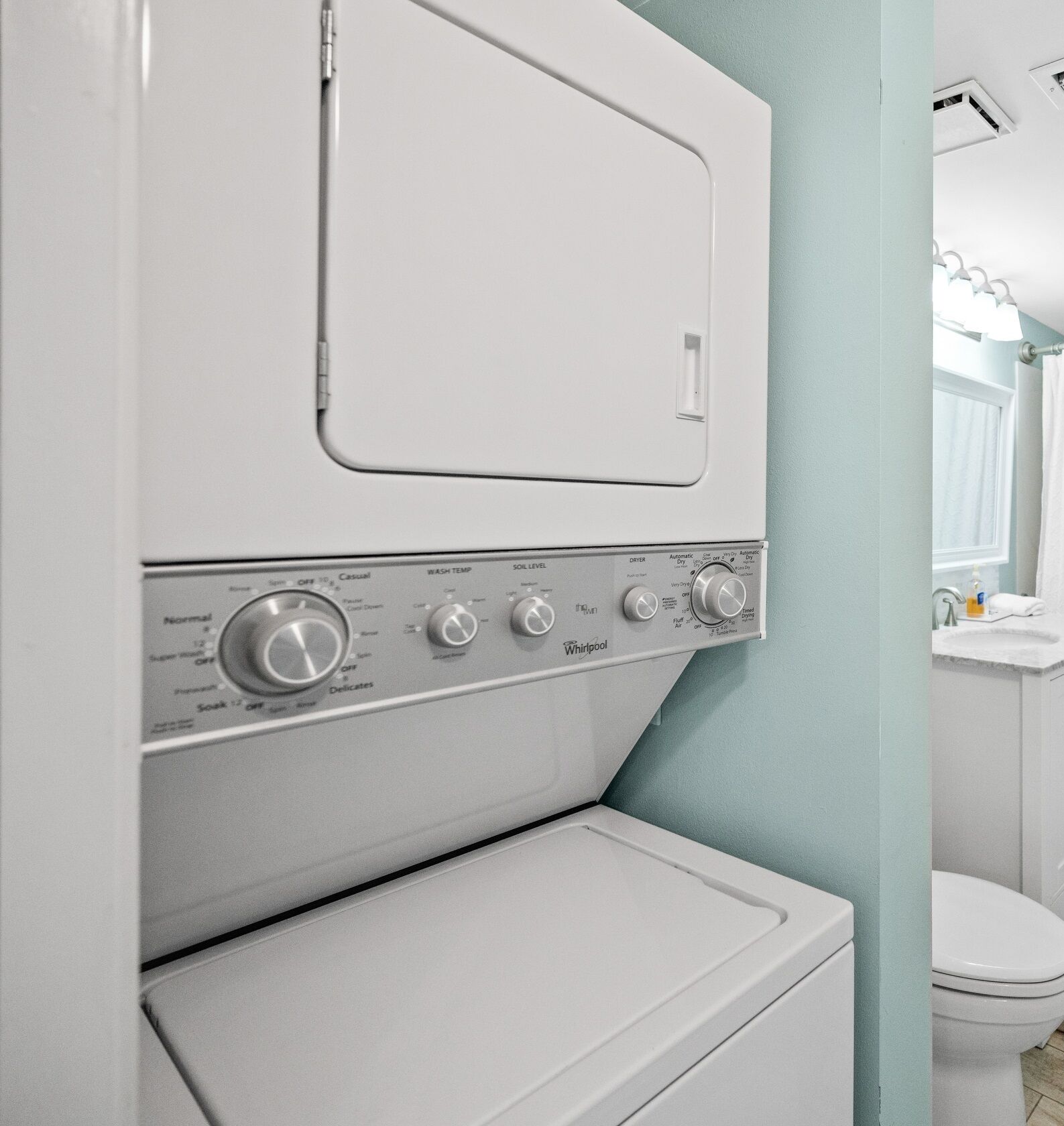 Eine Frontlader-Waschmaschine und -Trockner-Kombination in einem Badezimmer mit hellblauer Wand und sichtbarer weißer Toilette im Hintergrund.
