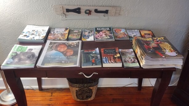 A table with various magazines and books, a basket, and a wall-mounted shelf with decorative items.