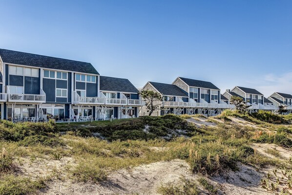 A row of modern beach houses with balconies, situated on a sandy dune with sparse vegetation.