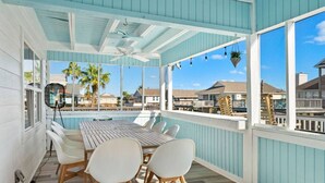 A covered outdoor dining area with a wooden table and white chairs, overlooking a beachfront with palm trees and houses. - Canal Front Home with Private Pool & Boat Slip (Galveston)