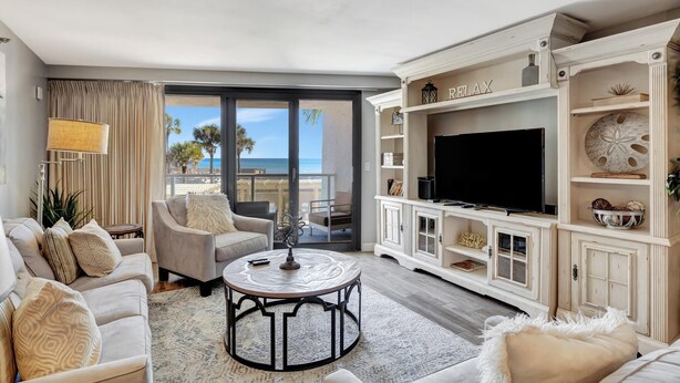 A spacious living room with a large TV, comfortable seating, and a balcony with a view of the ocean.