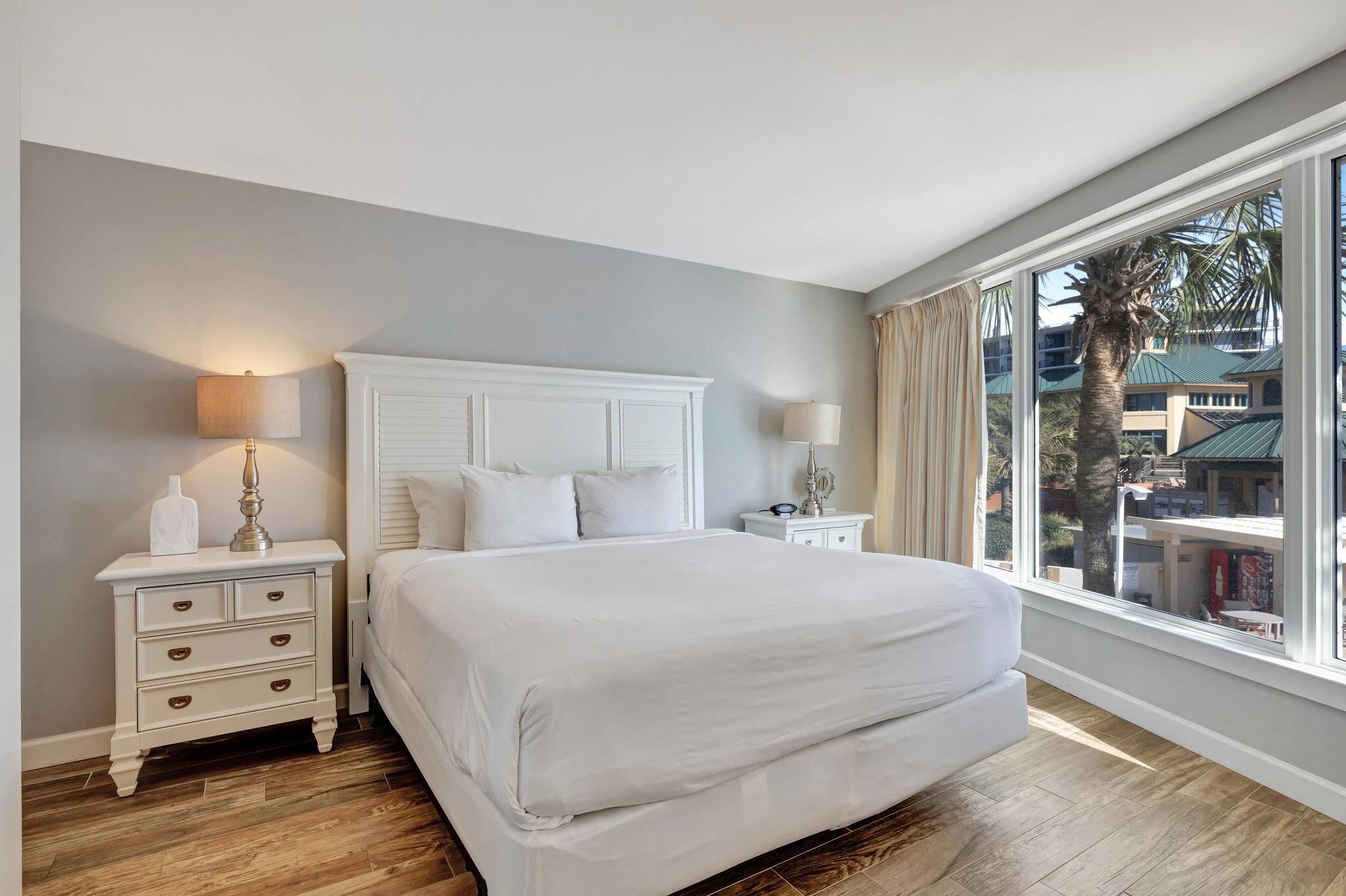 A bedroom with a large bed, white nightstands, and a view of palm trees and buildings through large windows.