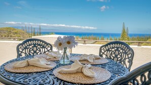 A round table with woven placemats and folded napkins, set against a scenic backdrop of a coastal landscape with trees and a clear sky. - KGV 24T8  Spacious 1BD Oceanview Villa (Lahaina)
