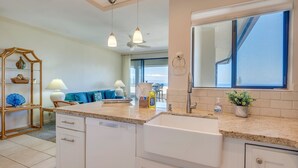 A modern kitchen with white cabinetry, a large sink, and a countertop with a mirror above it. There is a dining area visible through an open door, featuring a blue sofa and a table. - KGV 24T8  Spacious 1BD Oceanview Villa (Lahaina)
