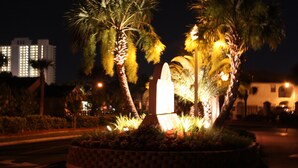 A nighttime view of a well-lit garden with a prominent obelisk, palm trees, and a building with illuminated windows in the background. - 153 Grand Island Blvd ~ Total Reno~Custom Kitchen~Granite~WiFi (Panama City Beach)