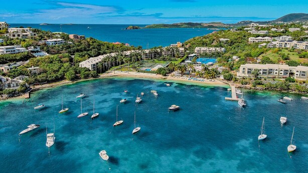 An aerial view of a coastal resort with multiple buildings, a marina with several boats, and a clear view of the sea and distant islands.