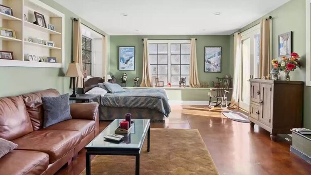 Ein geräumiges Wohnzimmer mit einem braunen Ledersofa, einem Glastisch und einem Bett mit grauer Bettdecke.