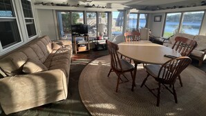Un salon spacieux avec une grande fenêtre donnant sur un lac, un canapé confortable, une table à manger avec des chaises et un téléviseur posé sur un meuble.