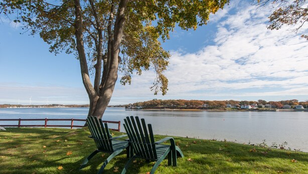 Deux fauteuils Adirondack sont placés sur une pelouse, avec un lac et des maisons en arrière-plan.