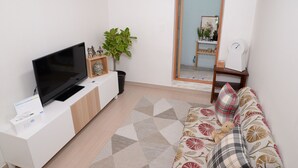 Ein Wohnzimmer mit einem Flachbildfernseher auf einer weißen Kommode, einer gepolsterten Couch mit gemusterten Kissen, einer Topfpflanze und einem Spiegel, der den Raum reflektiert.
