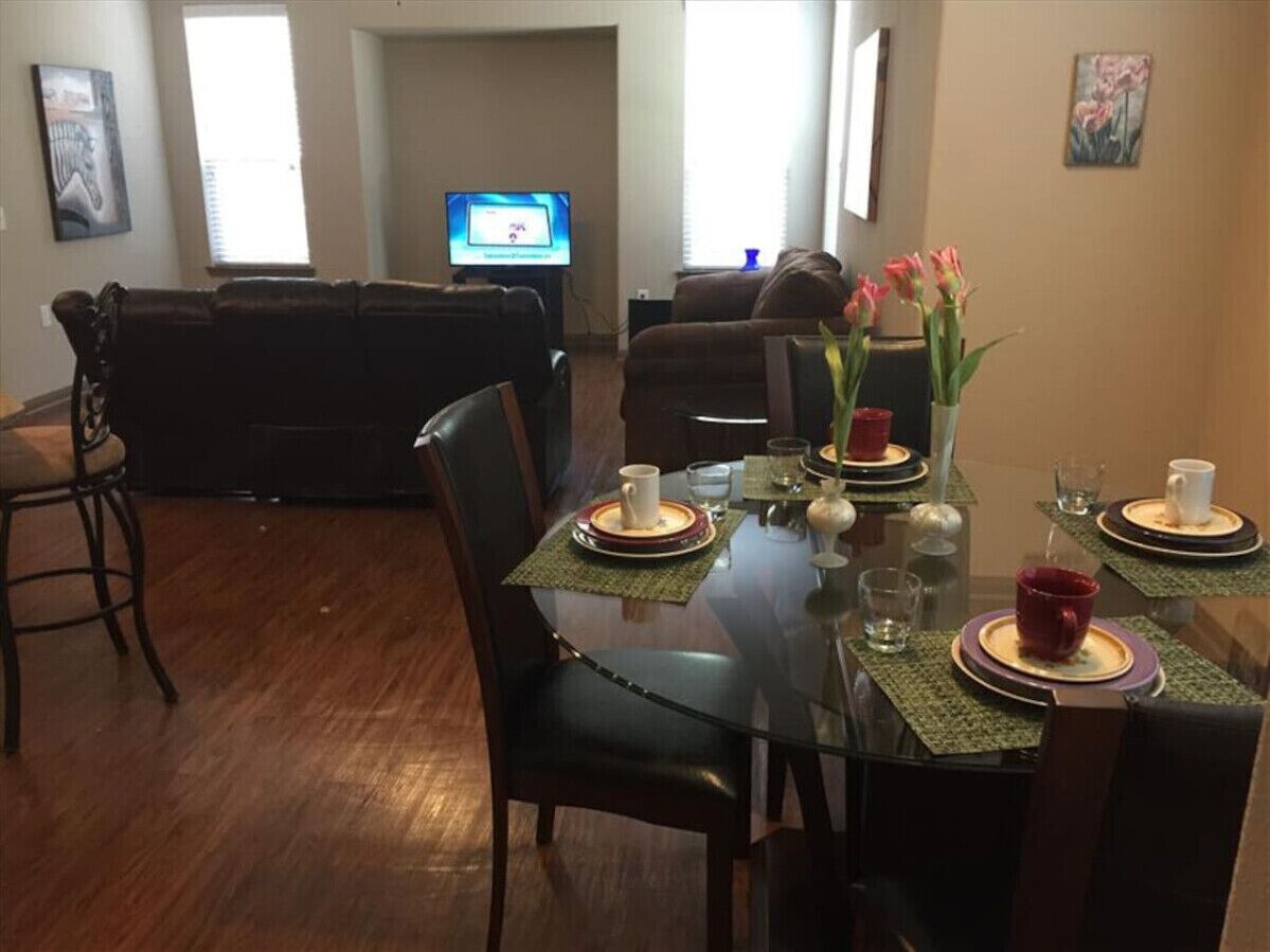 A dining area with a glass table set for four, surrounded by chairs. A television is mounted on the wall in the background.