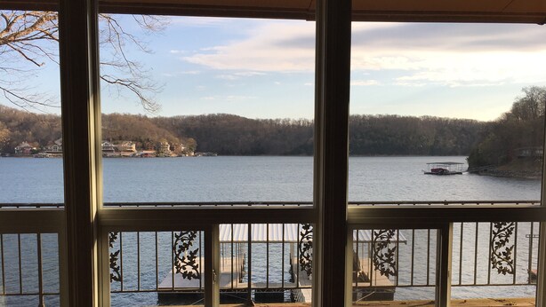 A view from a room with large windows overlooking a lake, with a dock and a boat visible in the distance. - 4 Bed and 3 Bath  - Lakefront with private dock (Camdenton)