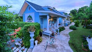 A blue cottage-style building with a porch, surrounded by lush greenery and a garden. - Little Cottage (Chanthaburi)