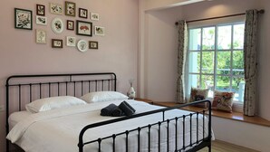 A bedroom with a bed, a window with curtains, and a wall with framed pictures. - Little Cottage (Chanthaburi)