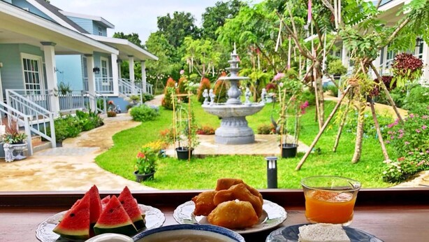 A dining table with a variety of dishes including watermelon slices, fried items, a bowl of soup, and a glass of orange juice, set against a view of a garden with a fountain and a house. - Little Cottage (Chanthaburi)
